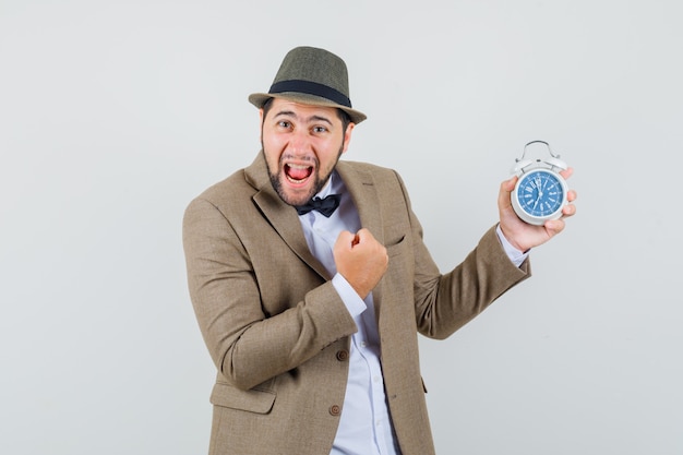 Free photo young man holding alarm clock with winner gesture in suit, hat and looking happy. front view.