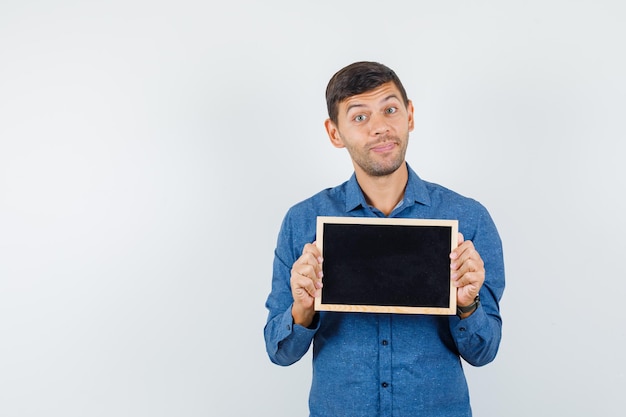 Young man holding blackboard in blue shirt and looking cheerful , front view.