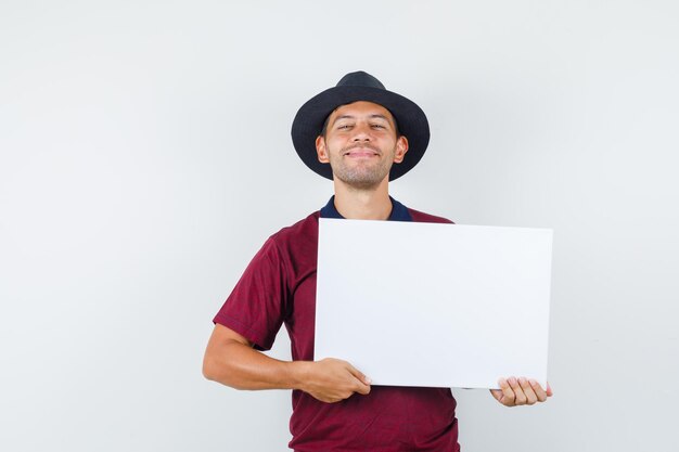 Young man holding blank canvas in t-shirt, hat and looking cheerful , front view.