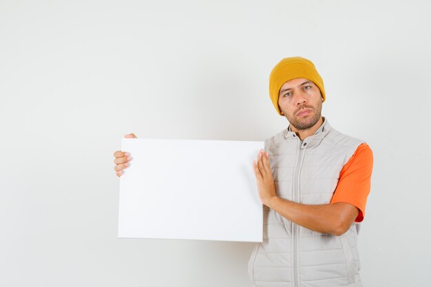 Young man holding blank canvas in t-shirt, jacket, hat and looking calm , front view.