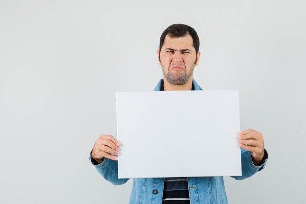 Young man holding blank canvas in t-shirt, jacket and looking discontent