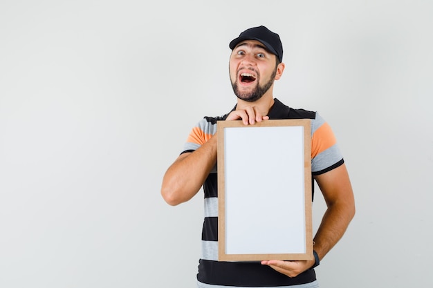 Young man holding blank frame in t-shirt, cap and looking happy , front view.