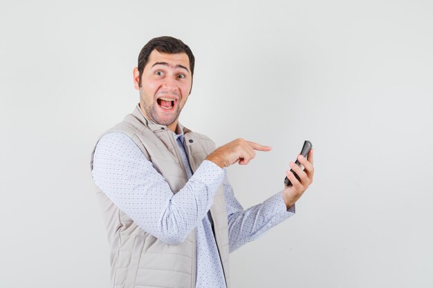 Young man holding calculator in one hand and pointing to it with index finger in beige jacket and cap and looking happy. front view.