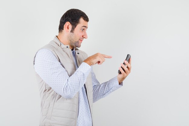 Young man holding calculator in one hand and pointing to it with index finger in beige jacket and cap and looking serious. front view.