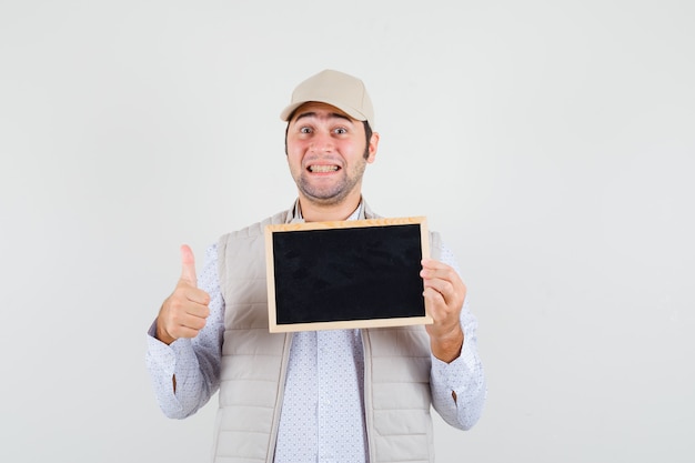 Young man holding chalkboard and showing thumb up in beige jacket and cap and looking happy , front view.