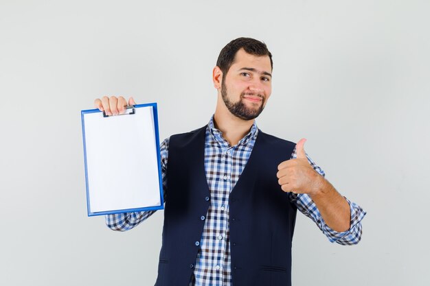 Young man holding clipboard with thumb up in shirt, vest and looking cheery , front view.