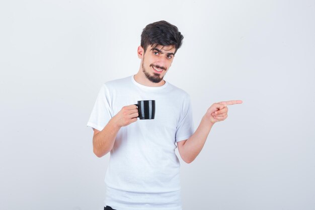 Young man holding cup of drink, pointing aside in white t-shirt and looking confident