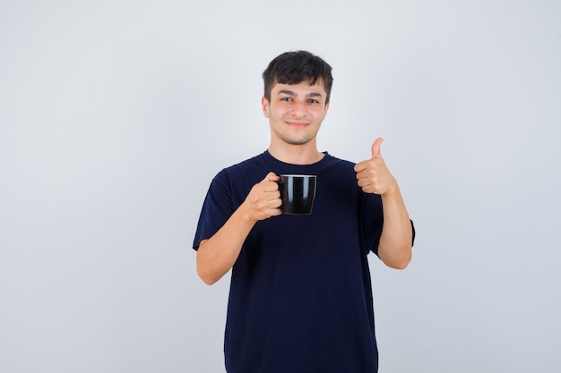 Young man holding cup of tea, showing thumb up in black t-shirt and looking cheery. front view.