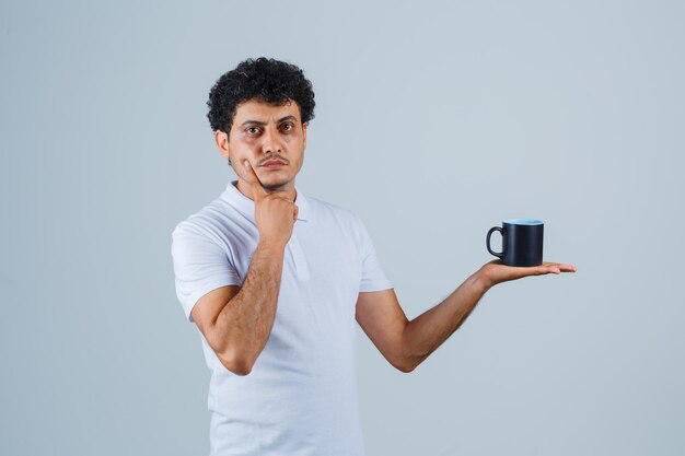 Young man holding cup of tea while leaning chin on hand in white t-shirt and jeans and looking pensive , front view.