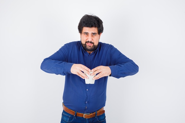 Free Photo young man holding cup with both hands in blue shirt and jeans and looking optimistic , front view.