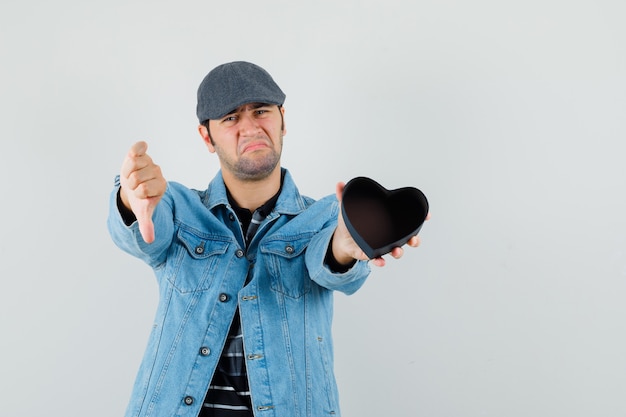 Free Photo young man holding empty gift while showing thumb down in jacket,cap and looking upset