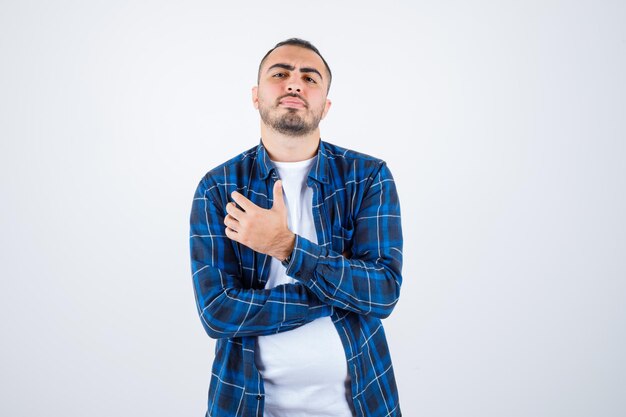 Young man holding hand on chest in checked shirt and white t-shirt and looking serious