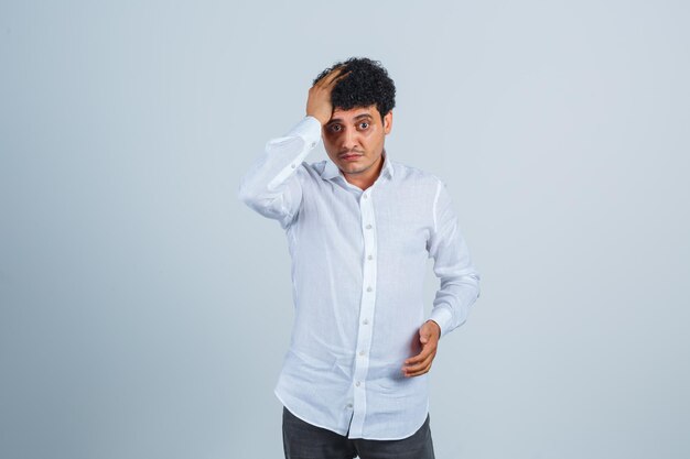 Young man holding hand on head in white shirt and looking wistful , front view.