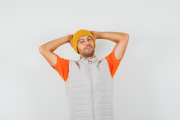 Free photo young man holding hands behind head in t-shirt, jacket, hat and looking relaxed. front view.