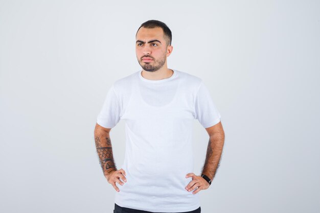 Young man holding hands on waist in white t-shirt and black pants and looking serious