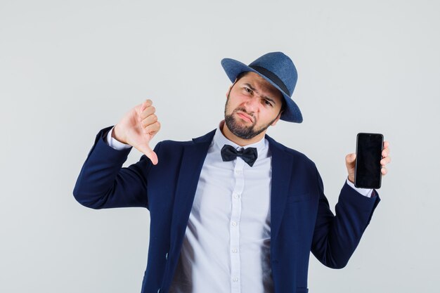 Young man holding mobile phone with thumb down in suit, hat and looking dissatisfied , front view.