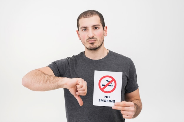 Free photo a young man holding no smoking sign showing thumbs down against white background