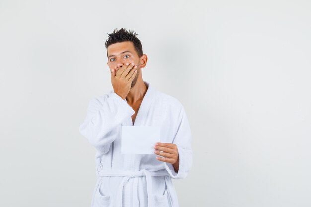 Young man holding paper sheet with hand on mouth in white bathrobe and looking surprised , front view.