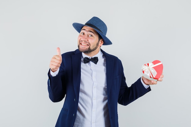 Young man holding present box with thumb up in suit, hat and looking happy , front view.