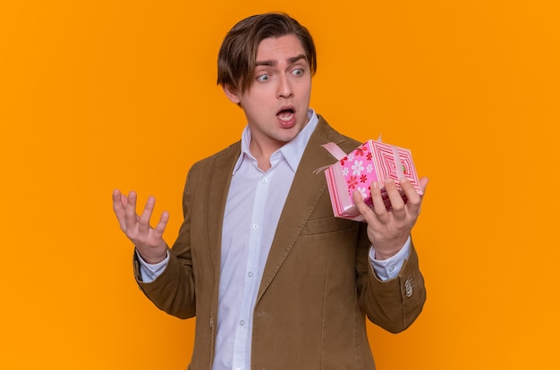 Young man holding present looking at it amazed and surprised going to congratulate with international women's day standing over orange wall