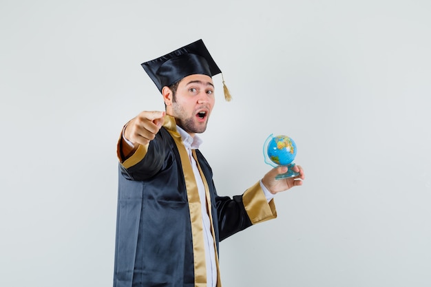 Young man holding school globe, pointing at camera in graduate uniform , front view.