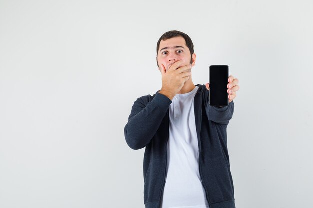 Young man holding smartphone, covering mouth with hand in white t-shirt and zip-front black hoodie and looking surprised. front view.