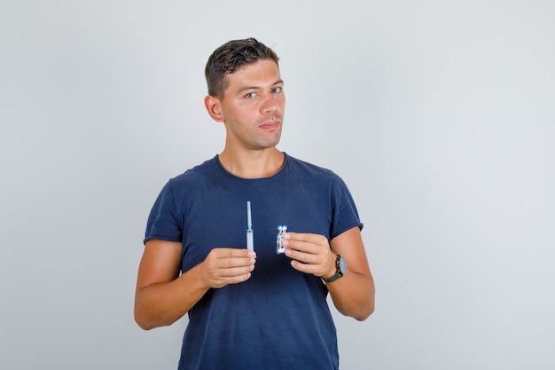 Young man holding syringe and vial in dark blue t-shirt and looking careful. front view.