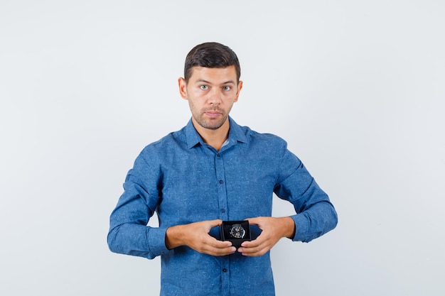 Young man holding watch box in blue shirt and looking sensible , front view.