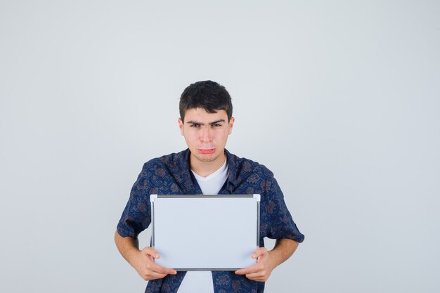 Young man holding whiteboard, curving lips in white t-shirt and floral shirt