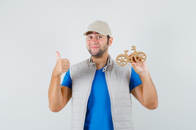 Young man holding wooden bike, showing thumb up in t-shirt, jacket and looking glad , front view.