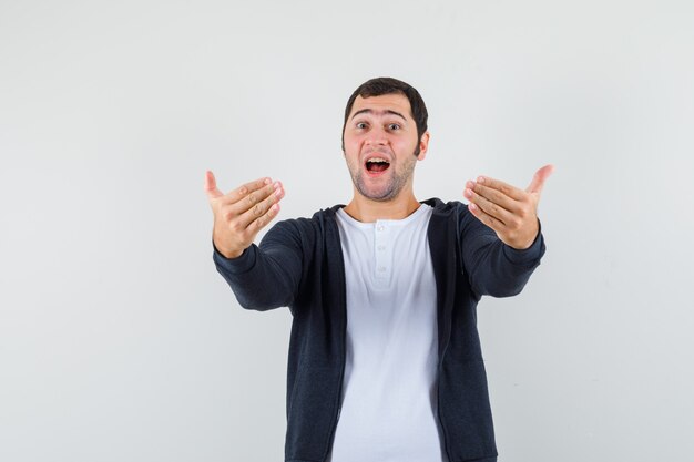 Young man inviting to come in white t-shirt and zip-front black hoodie and looking optimistic , front view.