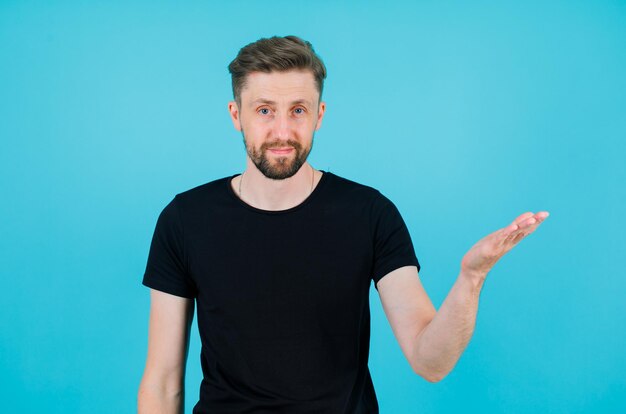 Young man is looking at camera by raising up his left hand on blue background