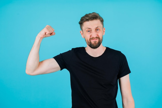 Young man is showing his muscle by raising up his fist on blue background