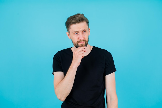 Free photo young man is thinking by holding hand on chin on blue background