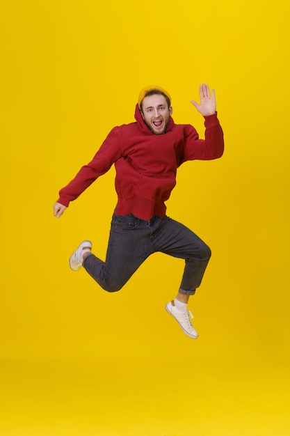 Free photo young man in joy jumping high wearing casual red hoodie and jeans isolated on yellow humorous hipster guy in jump