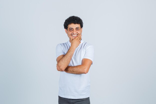 Young man keeping hand on chin in white t-shirt, pants and looking cheerful. front view.