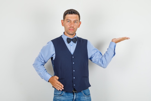 Young man keeping open palm with hand on waist in suit, jeans