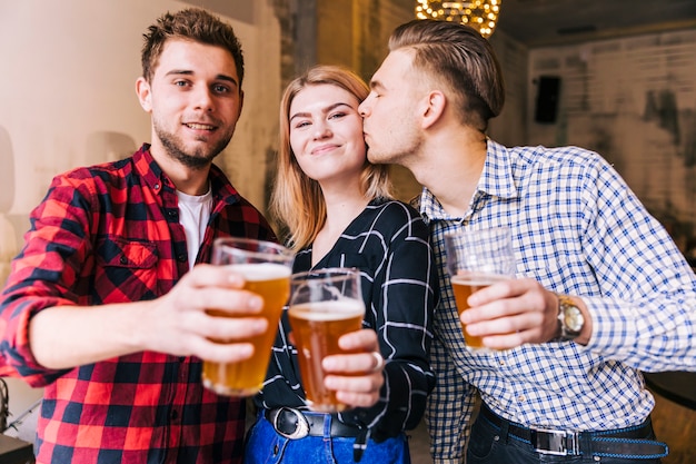 Young man kissing her girlfriend while toasting the beer glasses with friend