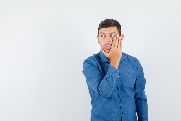 Young man looking aside with hand over cheek in blue shirt and looking scared , front view.