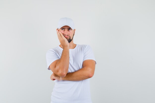 Young man looking away while propping cheek on hand in t-shirt,cap and looking serious. front view.