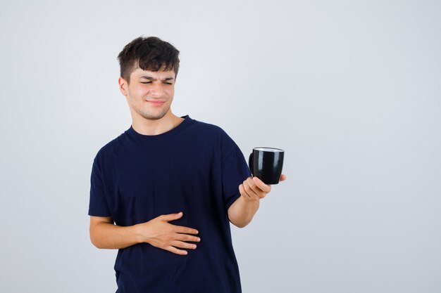 Young man looking at cup in black t-shirt and looking displeased , front view.