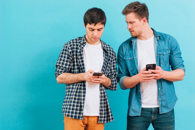 Young man looking at his friend texting on smartphone against blue backdrop