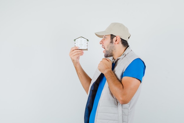 Free photo young man looking at house model in t-shirt, jacket, cap and looking amazed , front view.