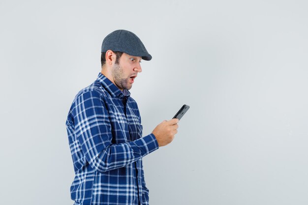 Young man looking at mobile phone in shirt, cap and looking surprised , front view.