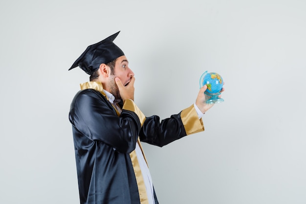 Free photo young man looking at school globe in graduate uniform and looking surprised .