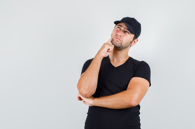 Young man looking up with finger on cheek in black t-shirt, cap and looking pensive