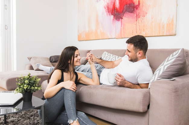 Young man lying on sofa holding hand of his girlfriend sitting on ground