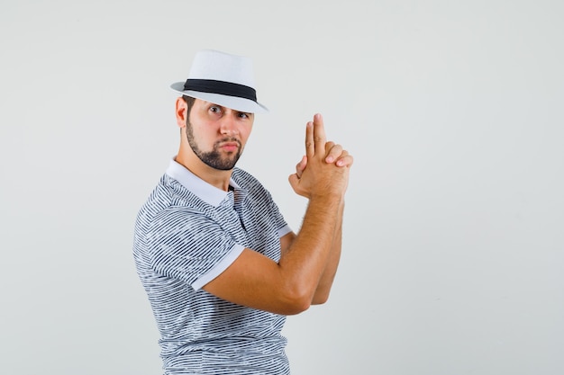 Young man making shooting gun gesture in striped t-shirt,hat and looking brave. front view.