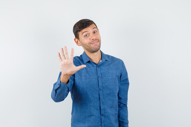 Young man making stop sign politely in blue shirt and looking jolly , front view.