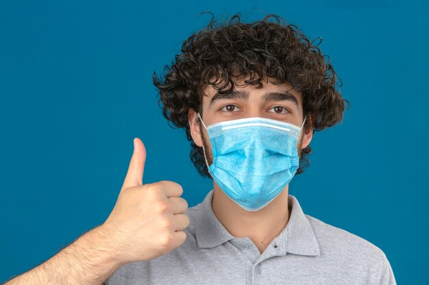 Young man in medical protective mask looking at camera with happy face showing thumb up over isolated blue background